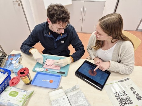 zwei Studierende üben Nähen von Wunden anhand des übermittelten Wissens über eine Video auf dem Tablet