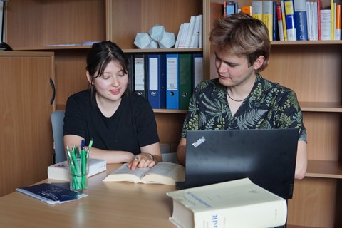 Foto zweier Studierender an einem Tisch. Vor den Studierenden liegen ein Laptop, drei Bücher und Stifte. Beide schauen gemeinsam in ein aufgeschlagenes Buch. 