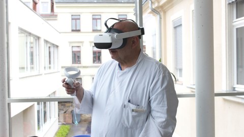 Farbfoto: Eine Person in weißem Kittel trägt eine VR-Brille und hält einen Joystick in der rechten Hand.