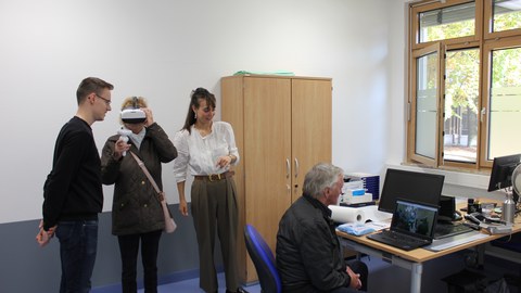 Foto:. Mittig steht eine Person mit einer VR-Brille auf dem Kopf und zwei Controllern in den Händen. Ihr links und rechts zugeneigt stehen zwei jüngere Personen. Rechts außen sitzt ein Mann und schaut auf den Bildschirm eines Laptops, der vor ihm steht.