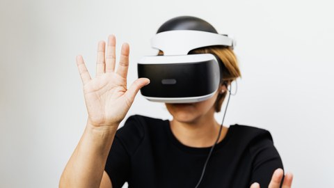 Farbfoto einer Person, die ein weißes VR-Headset trägt. Die Person hält ihre rechte Hand ausgestreckt vor ihr Gesicht.