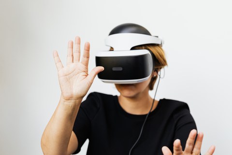 Farbfoto einer Person, die ein weißes VR-Headset trägt. Die Person hält ihre rechte Hand ausgestreckt vor ihr Gesicht.