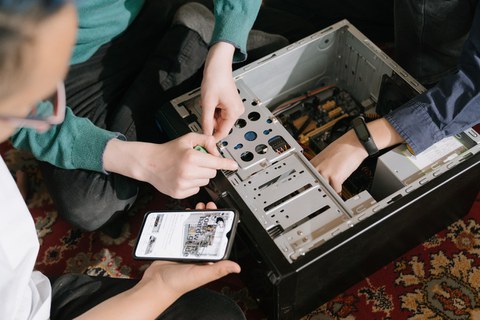 Farbfoto: Zwei paar Hände fassen in einen Kasten, in dem sich verschiedene Stromkabel und Leitungen befinden. Links davon hält eine Person ein Smartphon und schaut auf es herab.