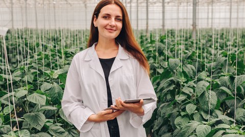 Eine Frau in einem weißen Laborkittel steht in einem Gewächshaus voller grüner Pflanzen. Sie hält ein Tablet in den Händen und lächelt, was auf Forschung oder Arbeit im Bereich Landwirtschaft oder Botanik hinweist.