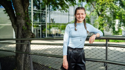 Eine junge Frau mit geflochtenen Zöpfen lehnt lächelnd an einem Geländer im Freien. Sie trägt ein hellblaues, langärmliges Oberteil und schwarze Shorts. Im Hintergrund sieht man ein modernes Gebäude mit Glasfronten und Bäume, die Schatten spenden.