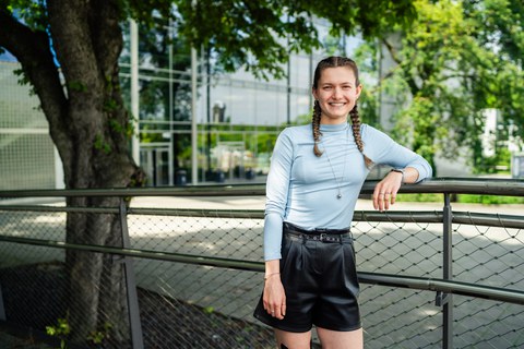 Eine junge Frau mit geflochtenen Zöpfen lehnt lächelnd an einem Geländer im Freien. Sie trägt ein hellblaues, langärmliges Oberteil und schwarze Shorts. Im Hintergrund sieht man ein modernes Gebäude mit Glasfronten und Bäume, die Schatten spenden.