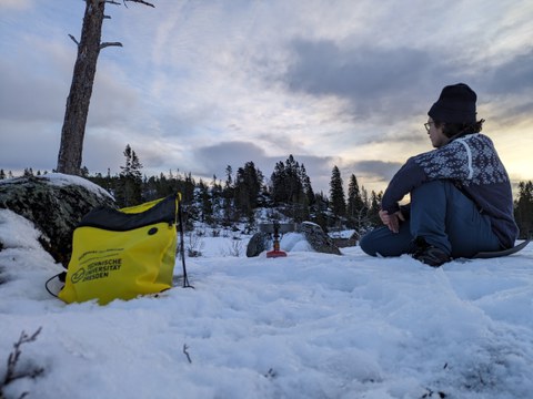  A person sits on the ground in a snow-covered winter landscape, facing a snowy forest in the background. Next to the person is a yellow bag with the logo of Technische Universität Dresden. 