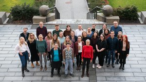 Gruppenfoto einer großen Gruppe von Menschen, die draußen auf einer Treppe steht. Sie blicken in die Kamera und lächeln. Im Hintergrund ist ein gepflasterter Weg, Grünflächen und Büsche zu sehen.