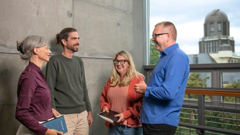 Four people stand in a bright building beside a concrete wall, smiling and talking together. Two of them are holding notebooks. Large windows in the background reveal a distinctive tower and some greenery outside. The atmosphere feels open, friendly, and collaborative.