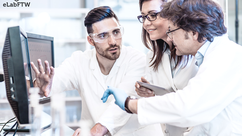 Three scientists in white lab coats collaborate in a modern laboratory. They are focused on a computer screen while one of them explains something. A female researcher holds a tablet, and the third scientist, wearing a blue glove, points at the screen. 