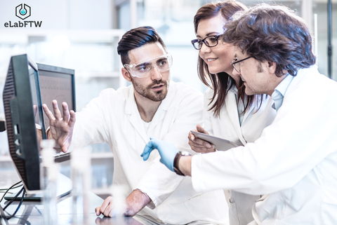 Three scientists in white lab coats collaborate in a modern laboratory. They are focused on a computer screen while one of them explains something. A female researcher holds a tablet, and the third scientist, wearing a blue glove, points at the screen. 