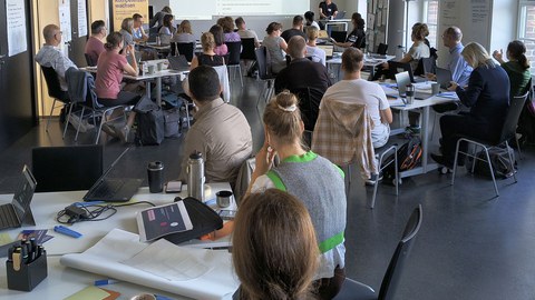 Teilnehmende der Lehrwerkstatt arbeiten in einem hellen Seminarraum an Tischen mit Laptops und Arbeitsmaterialien. Im vorderen Bereich sind Papier, Stifte und Moderationsmaterialien zu sehen. Im Hintergrund präsentiert eine Person Inhalte zu einer Lehrveranstaltung, die über einen Beamer projiziert werden. Die Gruppe diskutiert und arbeitet konzentriert an der Weiterentwicklung ihrer Lehre.