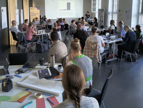 Teilnehmende der Lehrwerkstatt arbeiten in einem hellen Seminarraum an Tischen mit Laptops und Arbeitsmaterialien. Im vorderen Bereich sind Papier, Stifte und Moderationsmaterialien zu sehen. Im Hintergrund präsentiert eine Person Inhalte zu einer Lehrveranstaltung, die über einen Beamer projiziert werden. Die Gruppe diskutiert und arbeitet konzentriert an der Weiterentwicklung ihrer Lehre.