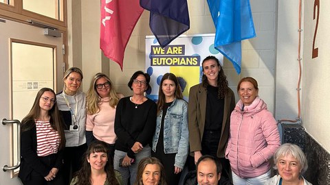 A group of around twelve people is standing and kneeling for a group photo indoors. Behind them hang three colorful flags and a poster reading “WE ARE EUTOPIANS.” The people are smiling at the camera.