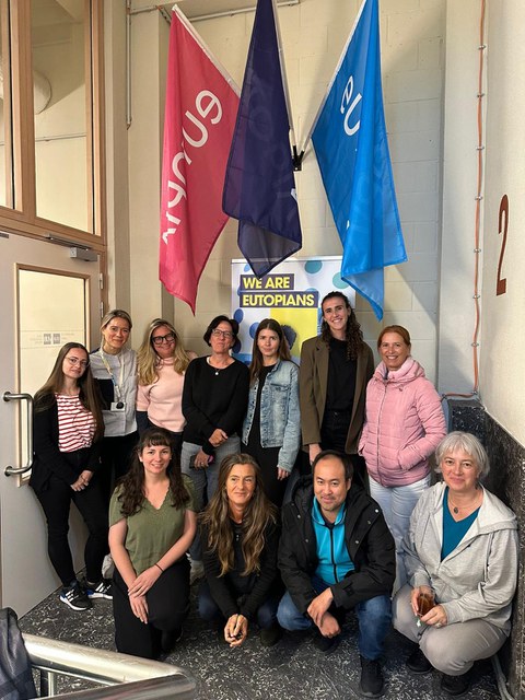 A group of around twelve people is standing and kneeling for a group photo indoors. Behind them hang three colorful flags and a poster reading “WE ARE EUTOPIANS.” The people are smiling at the camera.