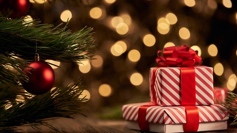 Two red-and-white striped Christmas presents with red ribbons placed next to a decorated pine branch, with warm, blurred lights in the background.