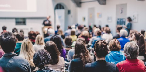 Blick in einen gut besuchten Vortragsraum: Viele Personen sitzen und hören einem unscharf im Hintergrund erkennbaren Vortragenden zu. Die Szene wirkt lebendig und konzentriert.