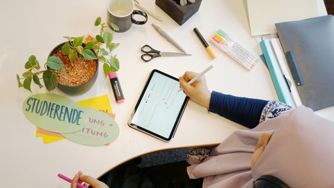 Aufsicht auf einen Schreibtisch: Eine Person schreibt mit einem Stift auf einem Tablet. Auf dem Tisch liegen Stifte, Schere, Marker, Notizkarten mit der Aufschrift „Studierende“, ein Becher sowie kleine Topfpflanzen.
