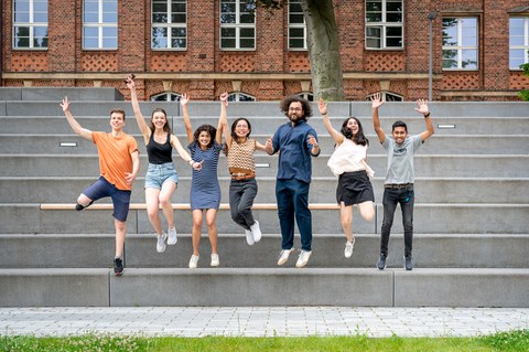 Eine Gruppe von sieben jungen Erwachsenen, die gemeinsam in die Luft springen, lächeln und jubeln. Sie stehen auf grauen Stufen vor einem Backsteingebäude mit großen Fenstern. Im Vordergrund ist eine Wiese zu sehen.