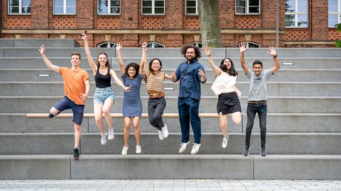 A group of seven young adults jumping in the air together, smiling and cheering. They are standing on gray steps in front of a brick building with large windows. A grassy area is visible in the foreground.