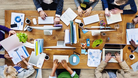 Das Foto zeigt einen großen Tisch aus der Vogelperspektive. Auf dem Tisch stehen Computer, Laptops und weitere Büromaterialien. Um den Tisch herum sitzen mehrer Personen und arbeiten an den Computern.