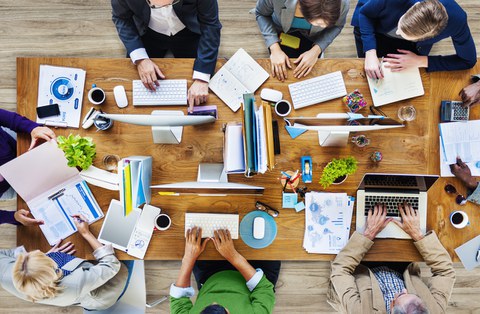 Das Foto zeigt einen großen Tisch aus der Vogelperspektive. Auf dem Tisch stehen Computer, Laptops und weitere Büromaterialien. Um den Tisch herum sitzen mehrer Personen und arbeiten an den Computern.