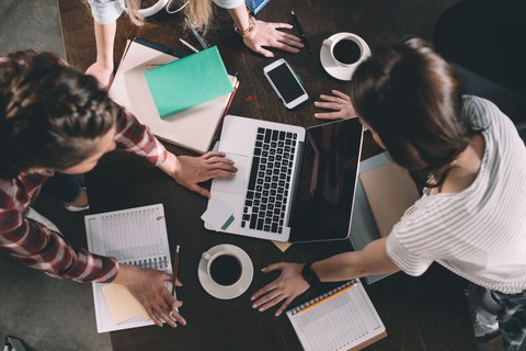 Three young people collaborate at a wooden table, surrounded by notebooks, a laptop, a smartphone, and coffee cups. They lean over their materials, with one person writing and another using the laptop. The scene conveys a productive and creative work.