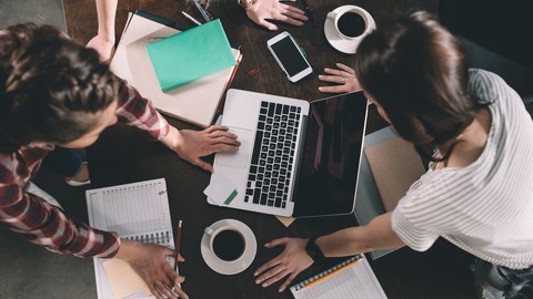 Three young people collaborate at a wooden table, surrounded by notebooks, a laptop, a smartphone, and coffee cups. They lean over their materials, with one person writing and another using the laptop. The scene conveys a productive and creative work.