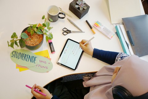 Foto einer Frau mit Kopftuch, die an einem Tisch sitzt und sich auf ihrem Tablet Notizen macht.