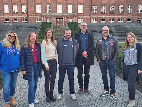 Group of eight adults standing side by side on a paved path in front of a large brick building with a staircase and symmetrical façade, all smiling at the camera.