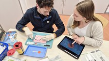 Zwei Studierende üben gemeinsam eine medizinische Nahttechnik an einem Trainingsmodell. Eine Person arbeitet praktisch, die andere unterstützt anhand einer digitalen Anleitung auf einem Tablet.
