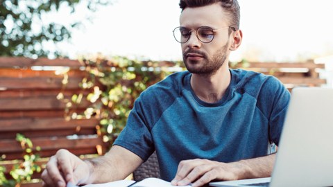 Das Foto zeigt einen Studenten an einem Tisch. Vor ihm steht ein Laptop und er schaut in sein Notizbuch.