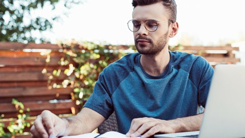 Das Foto zeigt einen Studenten an einem Tisch. Vor ihm steht ein Laptop und er schaut in sein Notizbuch.