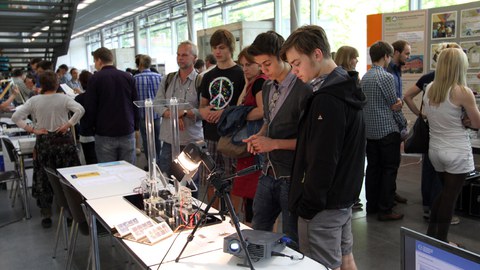 Das Foto zeigt einen Tisch mit technischen Geräten und einem Beamer. Vor dem Tisch stehen einige Studierende. Im Hintergrund erkennt man weitere Tische und Informationstafeln. Auch vor diesen stehen Studierende.