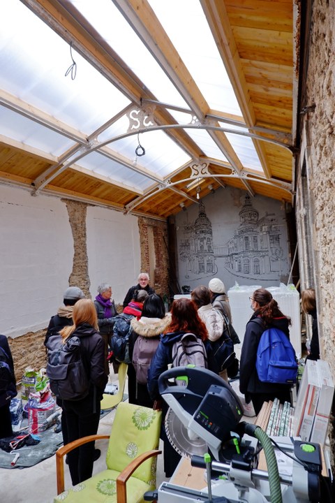 Zu sehen sind Personen in einem Gebäude mit einem Dach aus Holz und durchsichtigem Material. Die Wände sind aus Stein und nur an einigen Flächen verputzt. An der Front ist eine Zeichnung von historischen Gebäuden zu sehen.