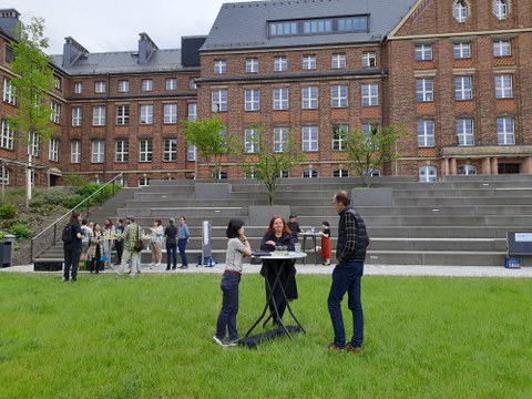 Zahlreiche Menschen stehen auf einer Wiese vor einem großen Gebäude und unterhalten sich.