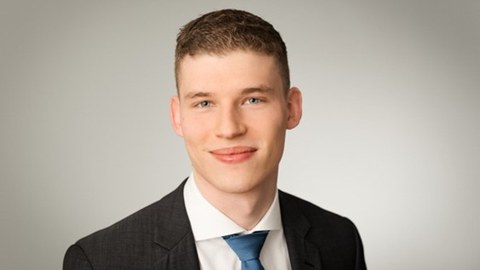Porträtfoto eines jungen Mannes mit kurzem braunem Haar, hellem Teint und blauen Augen. Er trägt einen dunklen Anzug, ein weißes Hemd und eine blaue Krawatte. Der Hintergrund ist neutral grau.
