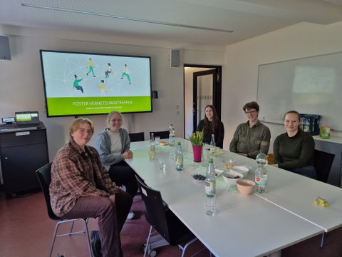 Fünf Personen sitzen an einem Tisch in einem Seminarraum während eines FOSTER-Vernetzungstreffens. Auf einem Bildschirm im Hintergrund ist eine Präsentationsfolie mit dem Titel „FOSTER Vernetzungstreffen – Vorstellung der Projekte von 2025“ zu sehen.