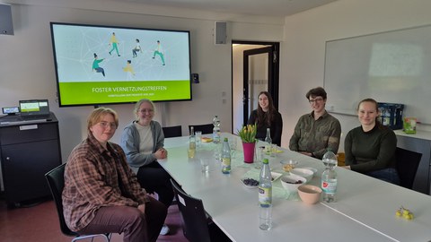 Fünf Personen sitzen an einem Tisch in einem Seminarraum während eines FOSTER-Vernetzungstreffens. Auf einem Bildschirm im Hintergrund ist eine Präsentationsfolie mit dem Titel „FOSTER Vernetzungstreffen – Vorstellung der Projekte von 2025“ zu sehen.