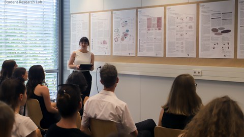 A woman stands in front of a pinboard and presents a poster. Other people sit in the audience and listen to her.