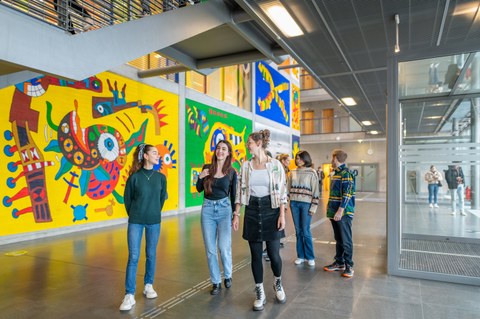 Eine Gruppe von jungen Leuten spaziert durch ein modernes Gebäude mit bunten, abstrakten Wandgemälden.