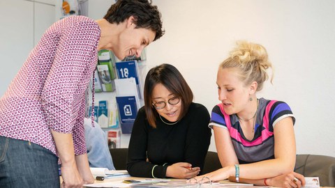 Two students are sitting at a table, bending over a file folder. A woman stands in front of them and looks at the documents.