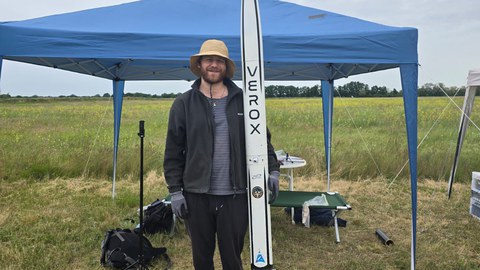 Eine Person mit Strohhut steht auf einer Wiese vor einem blauen Pavillon und hält eine große Modellrakete mit der Aufschrift „VEROX“. Im Hintergrund stehen Tische und Ausrüstung. Der Himmel ist bewölkt.