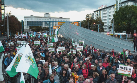  A large crowd at a demonstration in a German city. Many participants hold signs with slogans like “Arrest Habeck,” “Peace with Russia,” and “Out of NATO.” Some carry Saxon flags. Modern buildings and a tram stop are visible in the background.