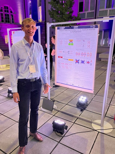 A young man in a light blue shirt and dark pants stands smiling next to a scientific poster. The poster is titled "Magnetic State Control of Non-van der Waals 2D Materials by Hydrogenation" and displays graphics and diagrams. The scene takes place outdoors at night, with the area illuminated by purple lighting. Other people and posters are visible in the background.