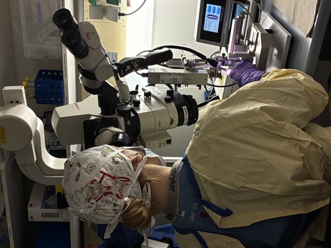 Person in Schutzkleidung arbeitet an einem Operationsmikroskop in einem Labor und führt einen präzisen mikrochirurgischen Eingriff durch.