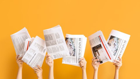 Four hands holding up different newspapers against a bright orange background. The newspapers display text and images related to business and finance.