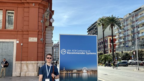 Person steht vor einem großen Plakat der „18th ACM Conference on Recommender Systems“ in Bari, Italien. Das Plakat zeigt das Datum der Konferenz (14.-18. Oktober 2024) und enthält das Logo der Veranstaltung. Im Hintergrund ist eine orangefarbene Fassade s