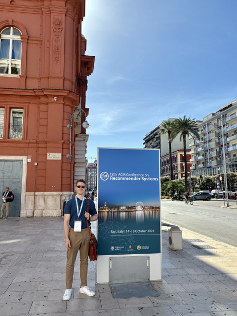 Person steht vor einem großen Plakat der „18th ACM Conference on Recommender Systems“ in Bari, Italien. Das Plakat zeigt das Datum der Konferenz (14.-18. Oktober 2024) und enthält das Logo der Veranstaltung. Im Hintergrund ist eine orangefarbene Fassade s