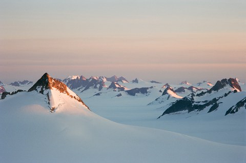 Schneebedeckte Berggipfel in weichem Abendlicht, die Spitzen leuchten rötlich in der tiefstehenden Sonne über einer weiten, weißen Gletscherfläche.